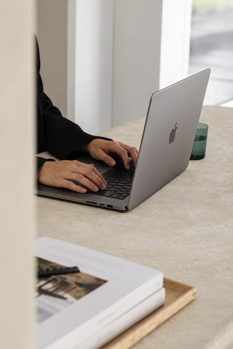 Mãos digitando em um notebook sobre a mesa, ao lado de um livro aberto