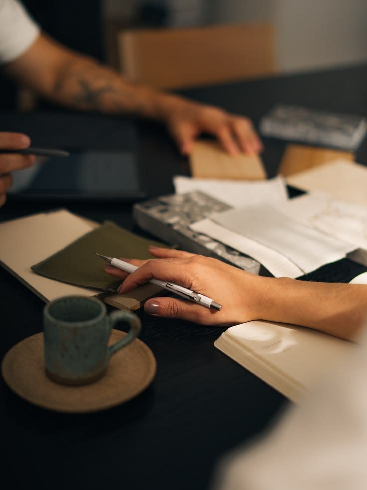 Mãos sobre uma mesa escura, com caderno, amostras de tecido e uma xícara de cerâmica