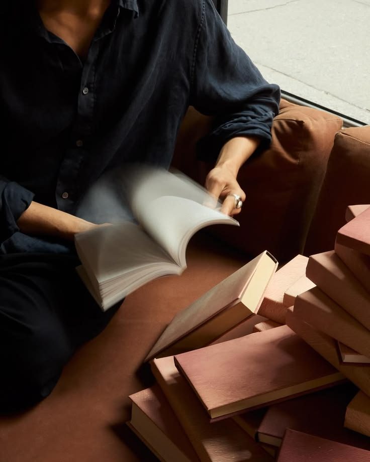 Pessoa de camisa escura folheando um livro, com uma pilha de livros rosados ao lado